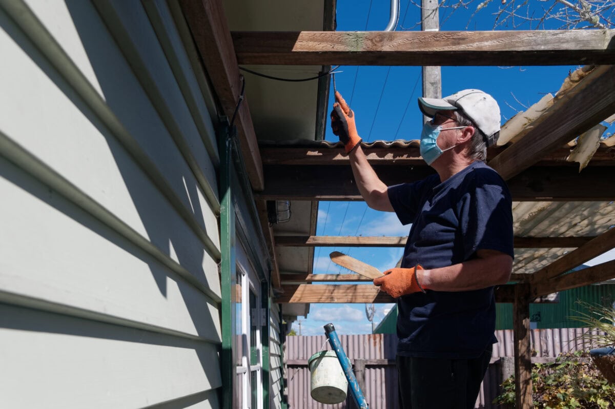 A worker or home handyman is up a ladder with a metal scrapper taking loose paint off a builder in preparation for painting