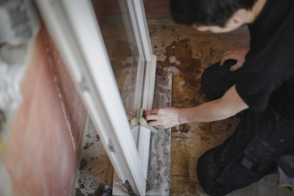 One handsome Caucasian young man with brown hair washes a window frame inside with a sponge and soap while sitting on his knees in a room being renovated, side view close-up with selective focus.